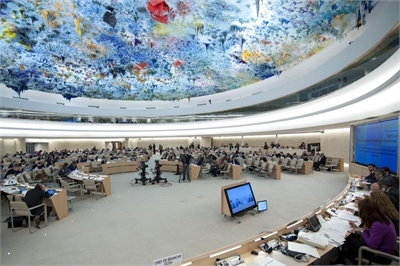 [ai] A large conference room filled with delegates seated at long tables, discussing matters. The ceiling features a colorful, artistic installation, and screens display information.